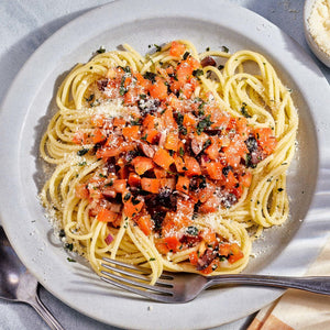 A plate of spaghetti topped with diced tomatoes, olives, red onion, and grated cheese, with a container of Gourmet Garden Basil (Case Of 72) placed beside it.