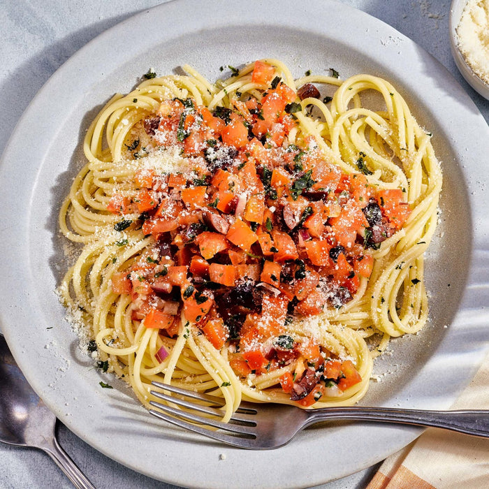 A plate of spaghetti topped with diced tomatoes, olives, red onion, and grated cheese, with a container of Gourmet Garden Basil (Case Of 72) placed beside it.