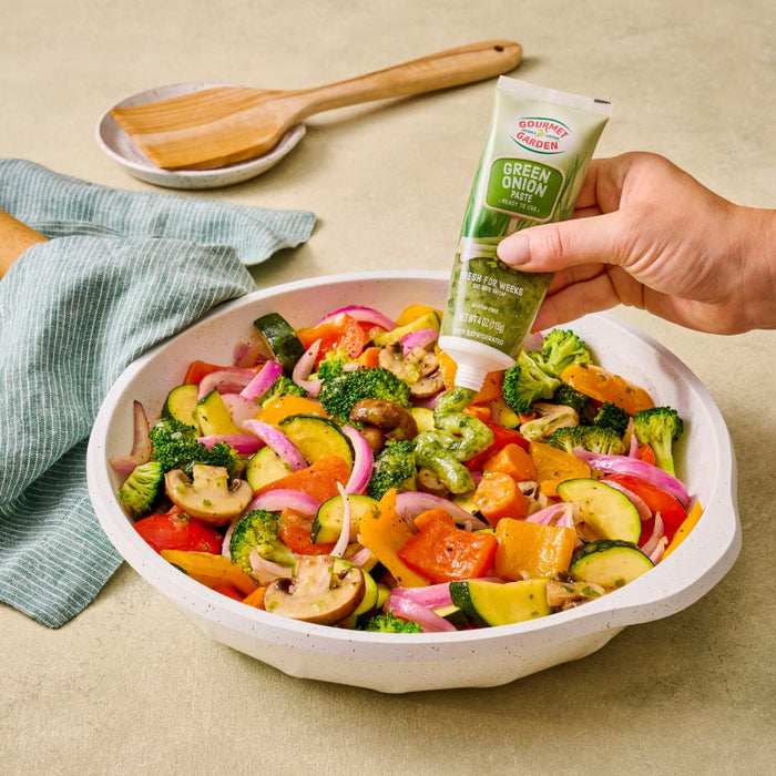 A person is adding Gourmet Garden basil paste to a bowl of colorful sauteed vegetables.