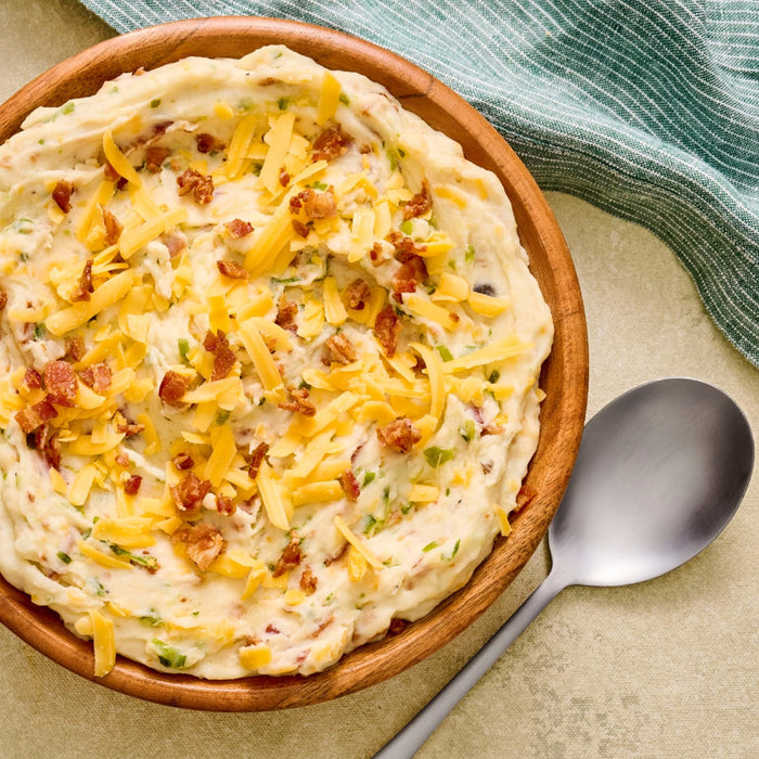 A bowl of creamy mashed potatoes topped with shredded cheese and bacon bits, served with a spoon.