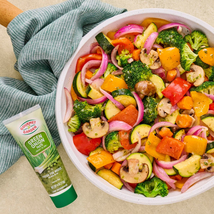 A colorful bowl of roasted vegetables, including broccoli, zucchini, mushrooms, red onion, and bell peppers, is displayed next to a tube of Amore Green Herb Paste.