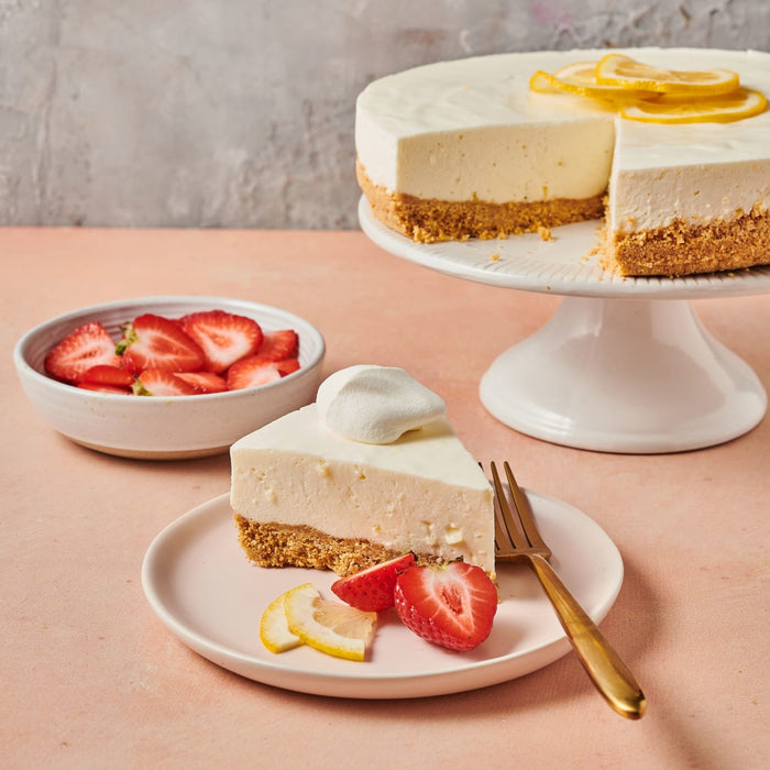 A slice of lemon cheesecake with whipped cream on top served with strawberries and lemon slices on a pink plate.