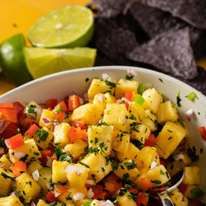 A bowl of pineapple salsa with red bell peppers red onion cilantro and a silver spoon.