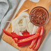 A wooden cutting board with sliced red peppers onions and a small bowl of red sauce.