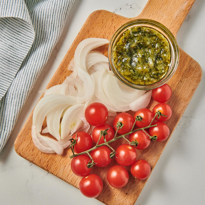 A wooden cutting board with sliced onions cherry tomatoes and a small bowl of green sauce.