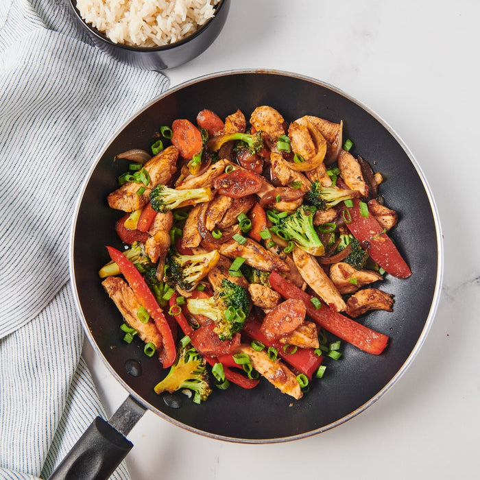 A black skillet with a handle is filled with a stir fry of chicken broccoli red peppers carrots and green onions.