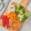 A wooden cutting board with a bowl of dip and various vegetables like carrots red peppers broccoli and onions.