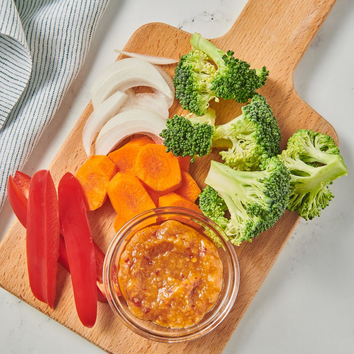 A wooden cutting board with a bowl of dip and various vegetables like carrots red peppers broccoli and onions.