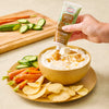 A hand squeezes Stonewall Kitchen Caramel Sauce from a tube onto a bowl of dip surrounded by chips, carrots, and cucumbers.