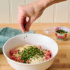 A hand sprinkles Gourmet Garden Italian Seasoning Blend into a bowl of ground meat and cheese.