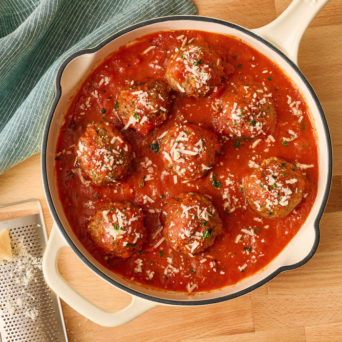 Overhead shot of meatballs in tomato sauce, sprinkled with parmesan cheese and herbs, served in a white skillet on a wooden surface with a grater and green cloth.