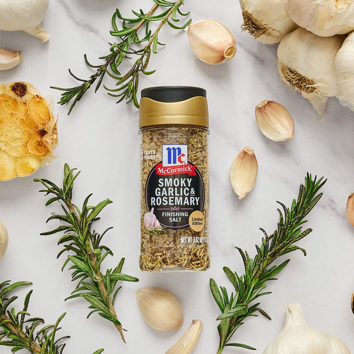 A bottle of McCormick Smoky Garlic &amp; Rosemary Finishing Salt is surrounded by garlic cloves and rosemary sprigs.