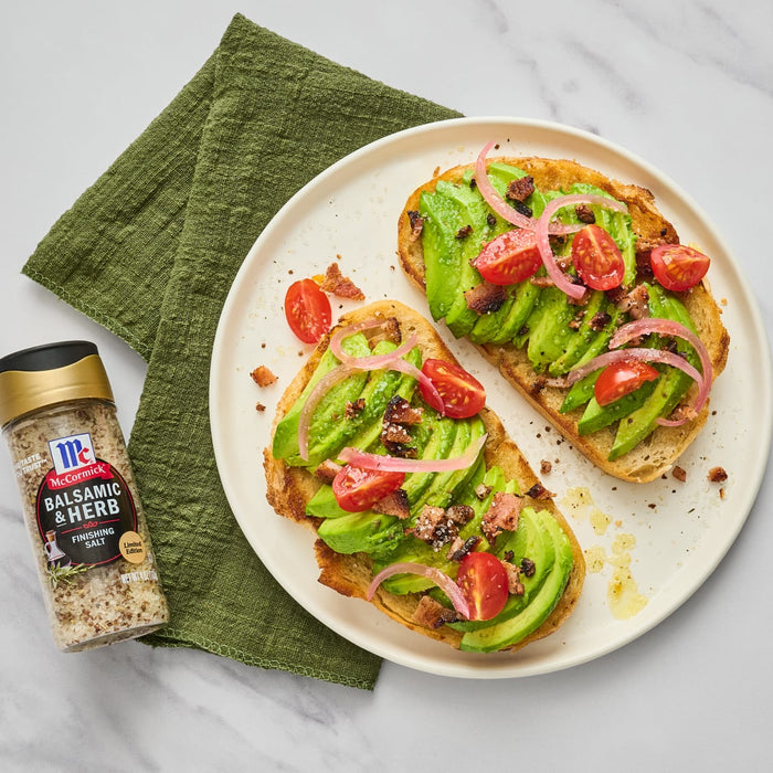 Two slices of avocado toast topped with bacon tomatoes red onion and McCormick Balsamic &amp; Herb Finishing Salt.