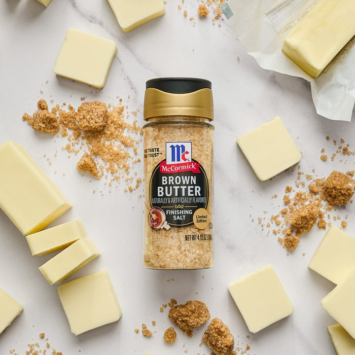 A jar of McCormick Brown Butter Finishing Salt sits on a white surface with pieces of butter and brown sugar.