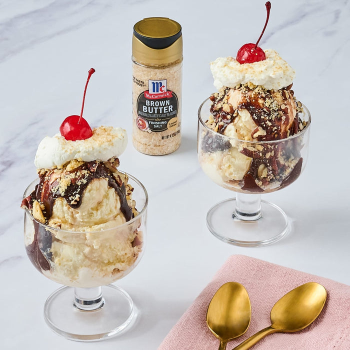 Two ice cream sundaes topped with whipped cream chocolate sauce and a cherry with a jar of McCormick Brown Butter Finishing Salt.