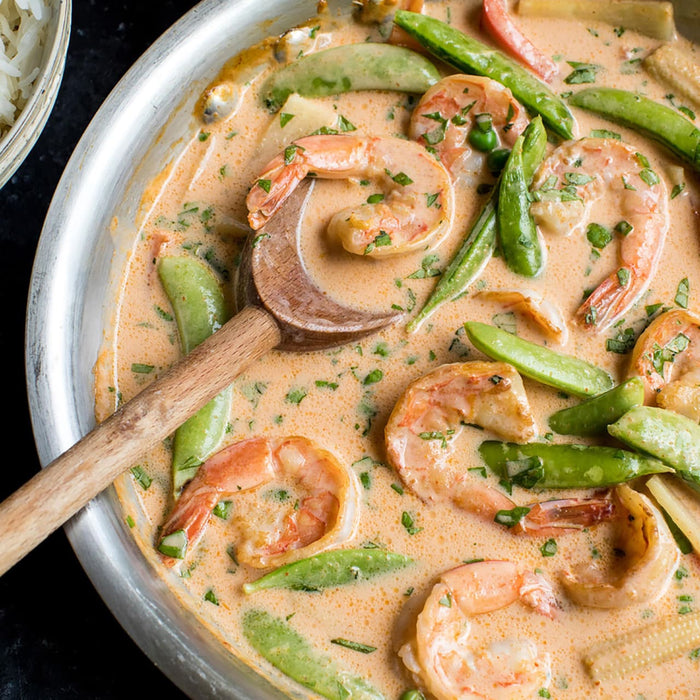 A pan of shrimp and snow pea curry is shown with a wooden spoon, highlighting a flavorful and visually appealing dish.