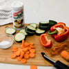 A cutting board with sliced cucumbers carrots and a red pepper a small bowl of salt a NoSalt salt substitute container and a vegetable peeler.