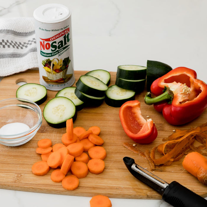 A cutting board with sliced cucumbers carrots and a red pepper a small bowl of salt a NoSalt salt substitute container and a vegetable peeler.