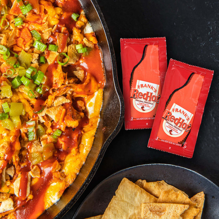 A cast iron skillet filled with a cheesy buffalo chicken dip with pita chips and two packets of Frank's RedHot sauce.
