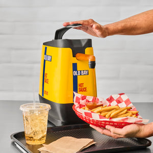 A hand pours Old Bay Hot Sauce onto french fries in a red basket.