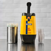 Yellow and black Old Bay hot sauce dispenser on a reflective surface with a gray tiled wall background.
