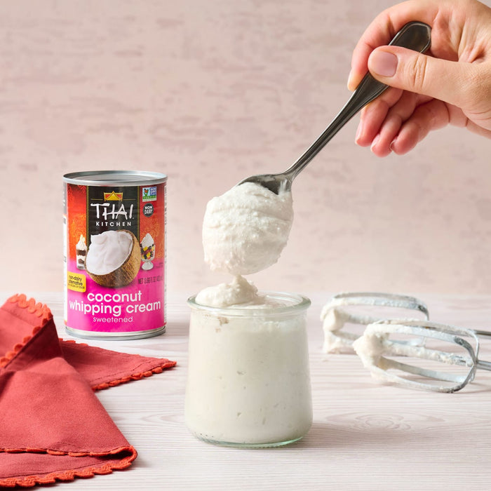 A can of Thai Kitchen coconut whipping cream is shown with a hand holding a spoonful of the whipped cream over a jar next to a red napkin and beaters.