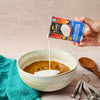 A can of Thai Kitchen evaporated coconut milk is being poured into a bowl of brown food.