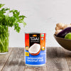 A can of Thai Kitchen evaporated coconut milk sits on a wooden table with fresh herbs and vegetables.