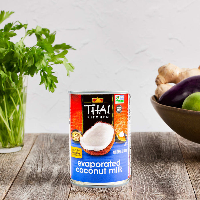A can of Thai Kitchen evaporated coconut milk sits on a wooden table with fresh herbs and vegetables.