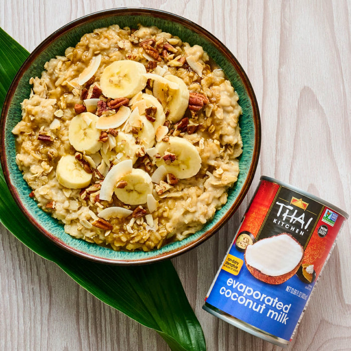 A bowl of oatmeal topped with banana slices coconut flakes and pecans is displayed next to a can of Thai Kitchen evaporated coconut milk.