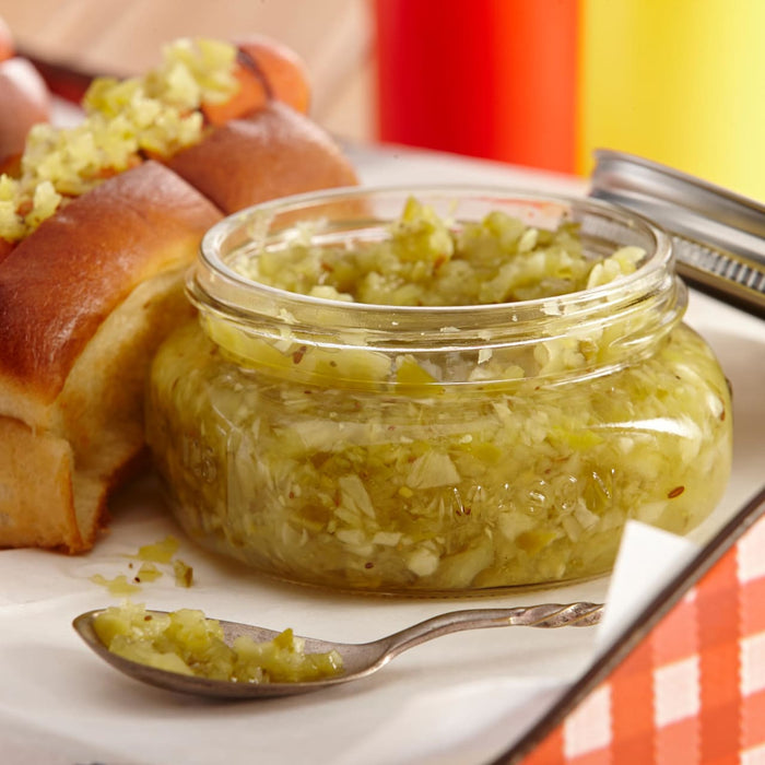 A jar of relish sits next to a hot dog bun with a spoon full of relish in the foreground.