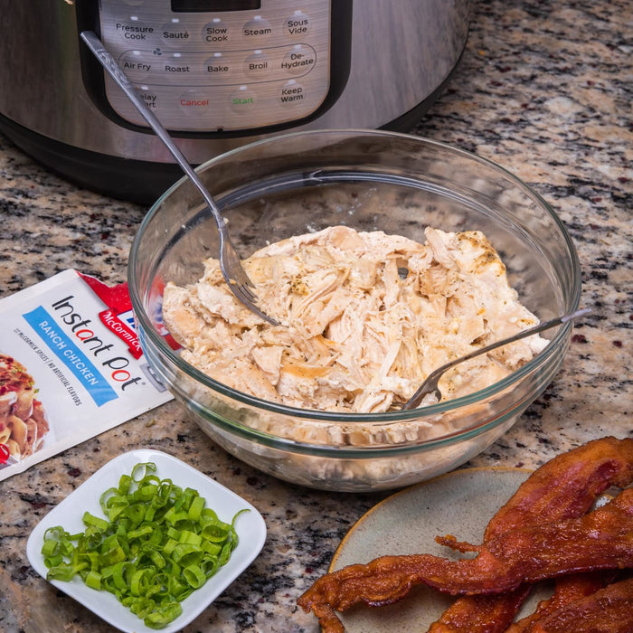 A bowl of shredded chicken with two forks a package of McCormick Instant Pot Ranch Chicken seasoning a plate of chopped green onions and a plate of cooked bacon.