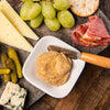 A white bowl with a dollop of mustard and a wooden handled knife surrounded by cheese grapes pickles and meat.