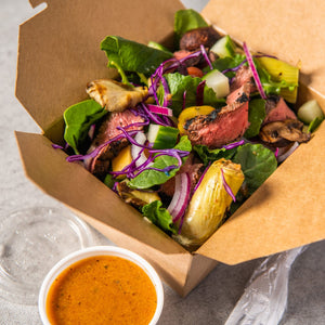 A close-up of a salad with grilled steak mushrooms and artichoke hearts in a to-go container with a side of dressing.