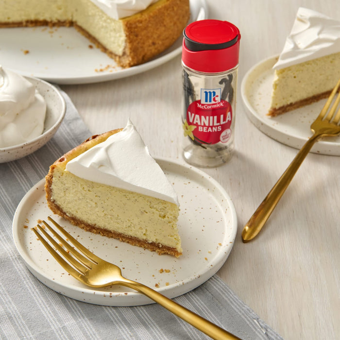 A slice of cheesecake with whipped cream on top, a jar of McCormick Vanilla Beans, and a fork on a white table.