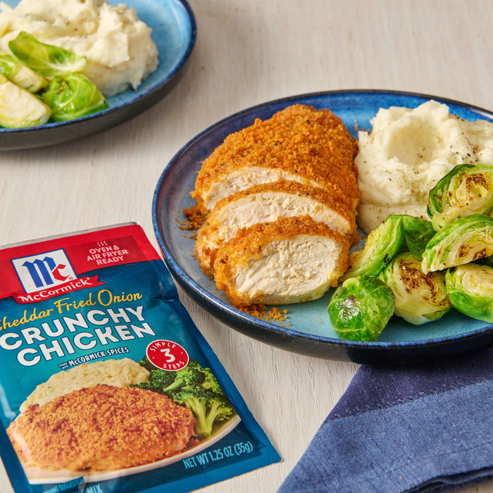 McCormick Cheddar Fried Onion Crunchy Chicken seasoning packet with a plate of breaded chicken, mashed potatoes and brussel sprouts.