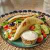 A plate of food with a pita bread sandwich, cucumber, tomato, and a side of dip.