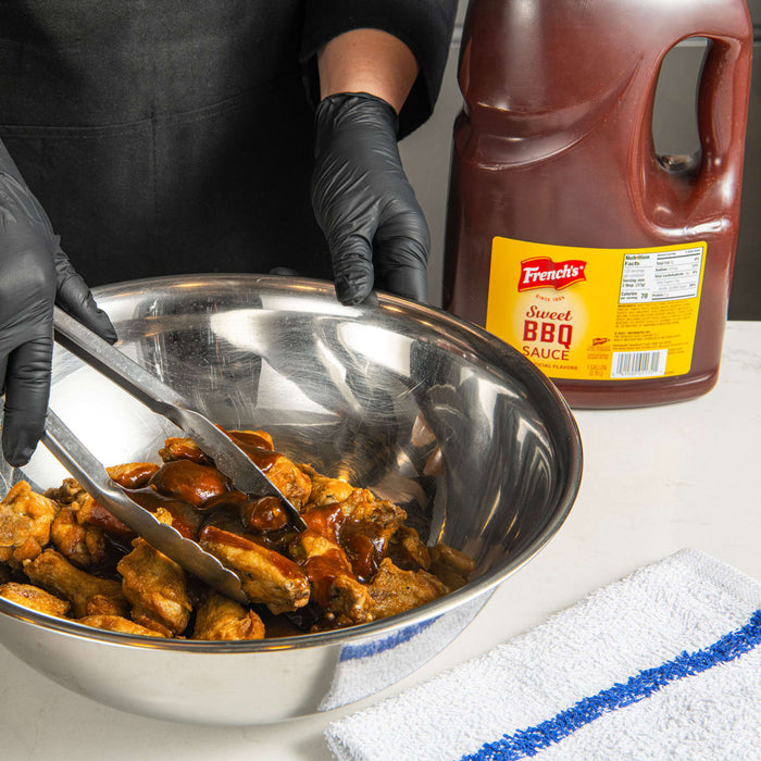 A person is using tongs to toss chicken wings in a bowl of French's Sweet BBQ Sauce.