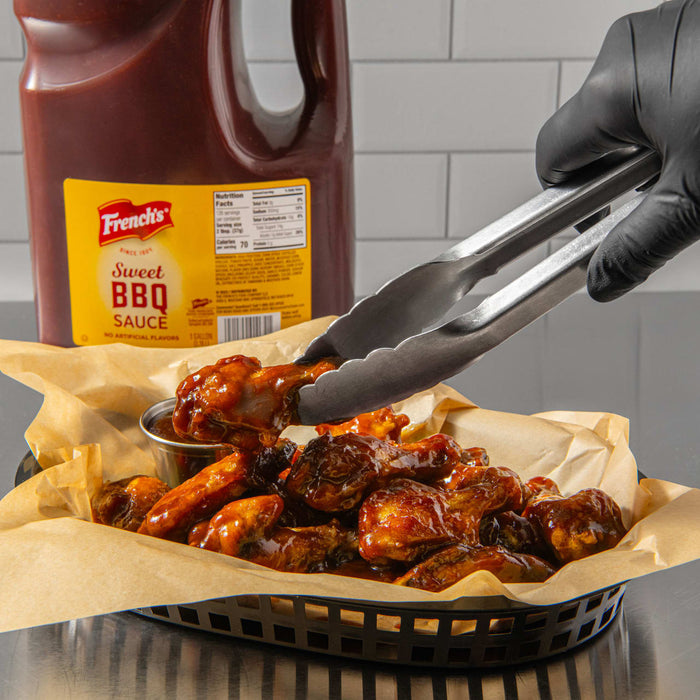 A hand in a black glove uses tongs to pick up a chicken wing from a basket of wings covered in French's Sweet BBQ Sauce.