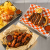 A metal tray with a burger, chips, and barbecue sauce, a tray with ribs, coleslaw, and barbecue sauce, and a tray with chicken wings and barbecue sauce.
