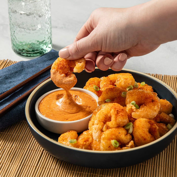 A hand dips a fried shrimp into a bowl of orange sauce, surrounded by other fried shrimp.