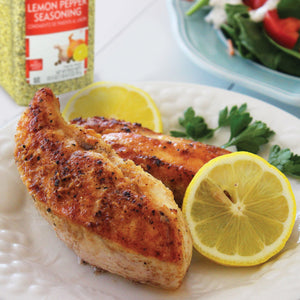 Two pieces of lemon pepper seasoned chicken breast with lemon wedges on a white plate.