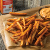 A close up of a pile of seasoned french fries on parchment paper next to a bowl of dipping sauce and a bottle of seasoning.