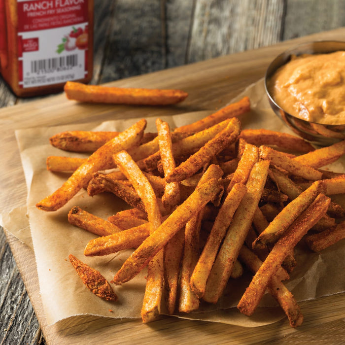 A close-up of a pile of crispy, seasoned french fries on parchment paper with a small bowl of ranch dressing in the background.