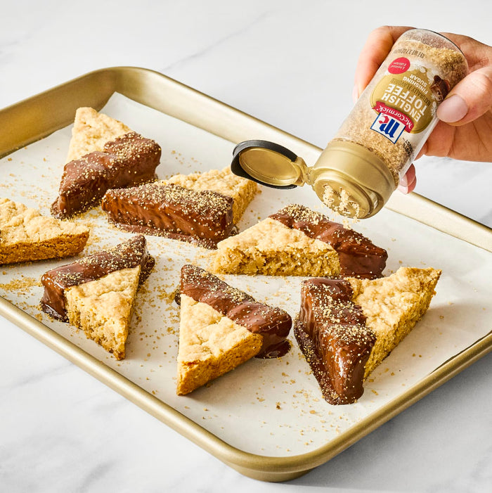 A hand sprinkles McCormick Cinnamon Sugar Blend on chocolate-dipped shortbread cookies on a baking sheet.