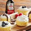Four mini cheesecakes topped with whipped cream and various berries next to a bottle of McCormick Pure Vanilla Extract.