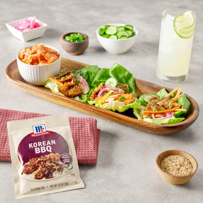 A packet of McCormick Korean BBQ seasoning mix is displayed next to a wooden tray of Korean BBQ lettuce wraps kimchi and other toppings.