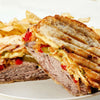 A grilled sandwich with sliced meat, cheese, and peppers is served with potato chips.