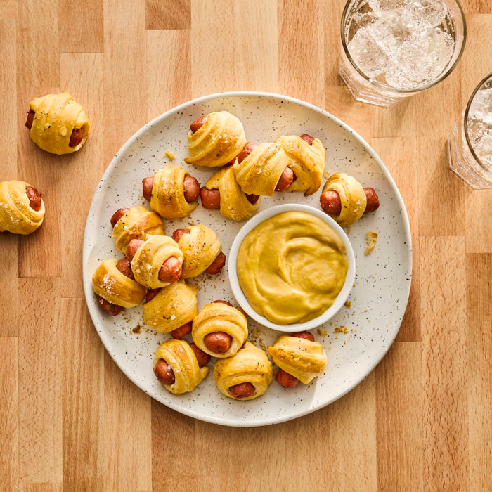 A plate of pigs in a blanket is served with mustard for dipping.