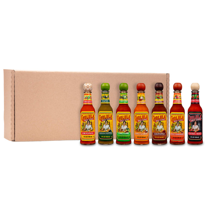 A lineup of seven different bottles of Cholula Hot Sauce, including Original, Green Pepper, Chili Lime, Chili Garlic, Chipotle, Sweet Habanero, and Extra Hot, are displayed in front of a cardboard box.